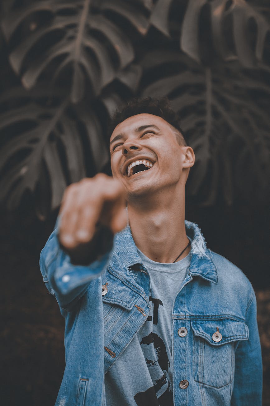 selective focus photo of man in blue denim jacket posing with his head back laughing while pointing finger